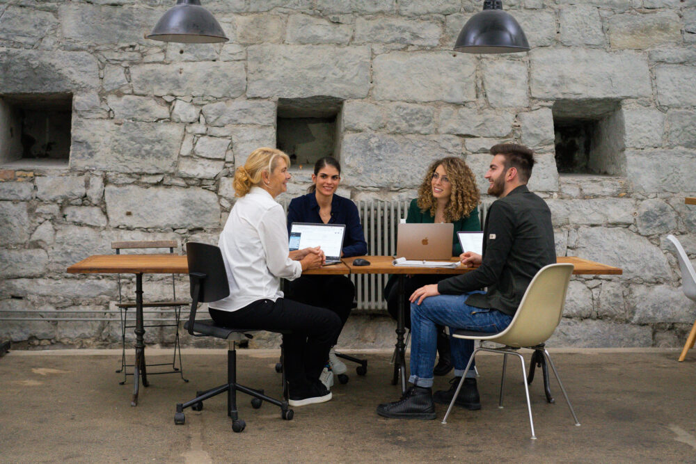 Travel Job Market während einer Teamsitzung im Grossraumbüro. Sie Sitzen miteinander an einem Tisch und diskutieren.